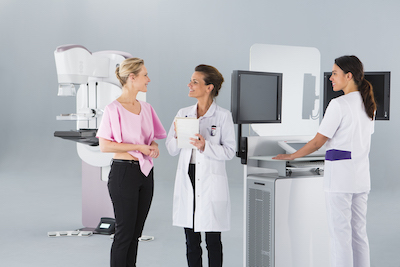 Picture of a female Radiology patient standing next to a Radiologist and a Radiologist Nurse. They are all smiling. There is a mammogram machine in the background.
