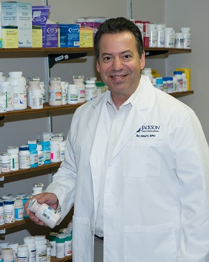 Picture of a smiling male Pharmacist holding a pill bottle while standing next to prescriptions.