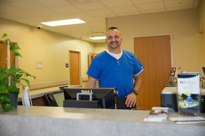 Picture of a male Nurse standing behind the Nurses Station, smiling.
