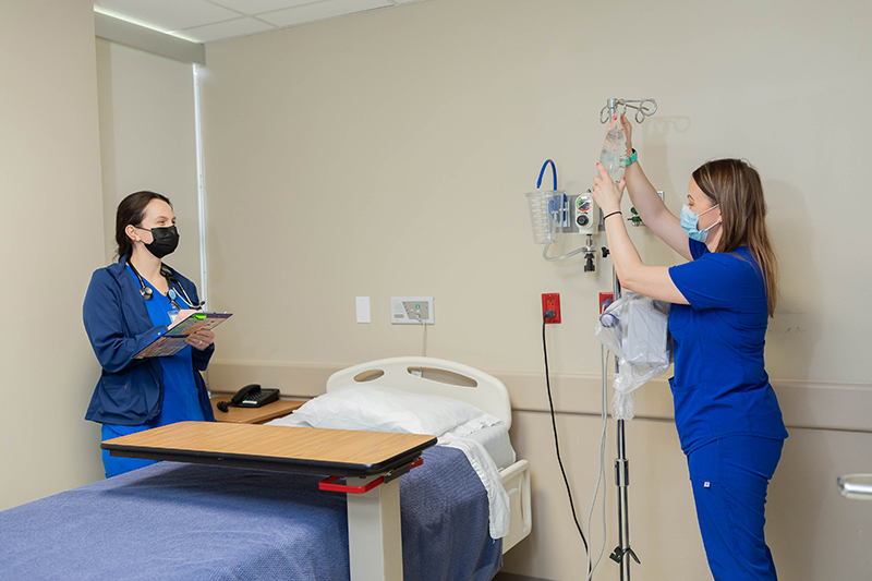 Picture of two females standing in a patient hospital room. One Nurse is standing next to a swing bed and holding a pen, clipboard, and paper and the other Nurse is taking an IV drip bag off of the IV pole.